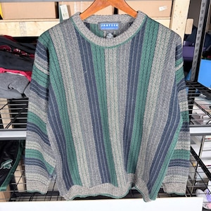 May include: A vintage crew neck sweater with vertical stripes in shades of green, gray, and navy blue. The sweater has a ribbed collar, cuffs, and hem. The brand name "Jantzen" is visible on a tag at the back of the neck.