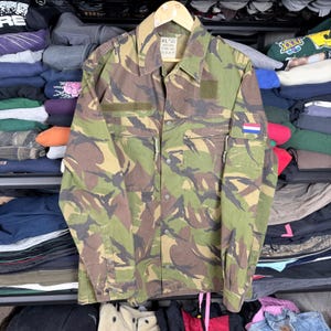 Puede incluir: Chaqueta militar de camuflaje en tonos verdes, marrones y negros. La chaqueta tiene múltiples bolsillos, un cuello y un parche con la bandera holandesa. Cuelga de una percha de madera.