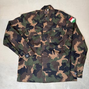 May include: A long-sleeve camouflage jacket featuring a green, brown, and black pattern. The jacket includes two chest pockets, a collar, buttoned cuffs, and a patch on the left shoulder with the Hungarian flag. A name tag reads HUNGARY.