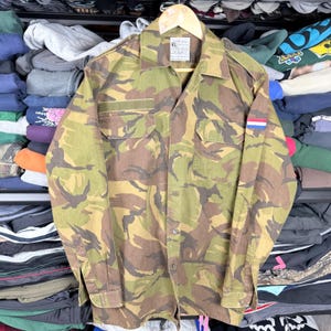 Puede incluir: Camisa de camuflaje con un parche de la bandera holandesa en la manga. La camisa presenta un patrón verde, marrón y beige, dos bolsillos en el pecho y un cuello. La camisa está colgada en una percha de madera.