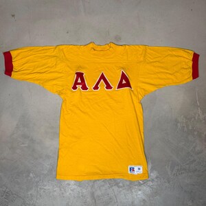 May include: A yellow short-sleeve jersey with red sleeve cuffs and the Greek letters Alpha, Lambda, and Delta in red with white outlines across the chest. A white Russell Athletic brand tag with the size M is visible at the bottom left corner.