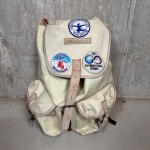 May include: An off-white canvas backpack featuring a large top flap with three circular embroidered patches and a rectangular patch labeled Lillehammer. The bag includes two front pockets and a central vertical leather strap with a metal buckle.