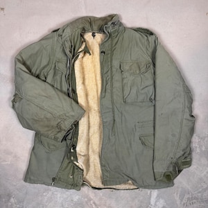 May include: An olive green field jacket featuring a high collar, a front zipper closure with a buttoned storm flap, and two large chest pockets. The interior is lined with a thick, beige, textured fleece material, providing insulation.