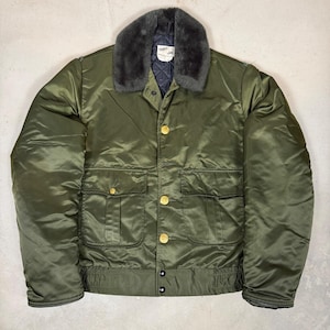 May include: An olive green bomber jacket featuring a dark gray faux fur collar, a quilted dark interior lining, and a front closure with gold-toned buttons. The jacket includes two large front pockets with button flaps and a label reading TUFFY JAC.