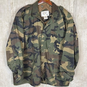 Può includere: Giacca con motivo mimetico nei toni del verde, marrone e nero. La giacca ha più tasche, una chiusura frontale con bottoni e un colletto. La giacca è appesa a una gruccia di legno.