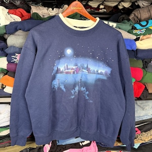 May include: Navy blue sweatshirt with a cream-colored neckline. The front features a nighttime landscape scene with a full moon, stars, a farm, and a body of water. The sweatshirt is made of a soft, comfortable material.