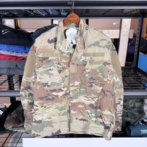 Puede incluir: Chaqueta militar con estampado de camuflaje con múltiples bolsillos y parches. La chaqueta está colgada de una percha de madera. Los colores incluyen tonos de verde, marrón y beige. La chaqueta tiene una parte delantera abotonada y un cuello.