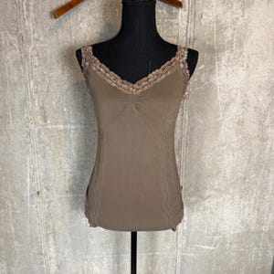May include: A taupe-colored camisole featuring a V-neckline trimmed with intricate floral lace detailing. The garment includes a gathered center bust detail and vertical ribbed texture panels along the sides, providing a fitted silhouette.