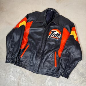 Pode incluir: Um casaco de couro preto com painéis vermelhos e amarelos nas mangas e no tronco. O peito apresenta um emblema circular com o texto Tide Racing Team em branco e laranja. O casaco inclui uma gola, fecho de correr frontal e dois bolsos.