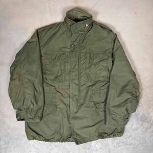 May include: An olive green field jacket featuring a high collar with a zip closure, a front button placket, and four large flap pockets on the chest and waist. The long sleeves have adjustable cuffs, and the shoulders include epaulettes.