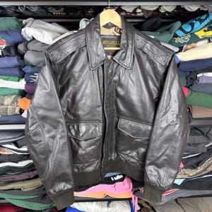 Könnte beinhalten: Eine braune Lederjacke Typ A-2 mit Kragen, zwei Vordertaschen und geripptem Saum und Bündchen. Die Jacke hängt an einem Holzbügel. Die Jacke hat ein Etikett mit der Aufschrift "JACKET TYPE A-2".