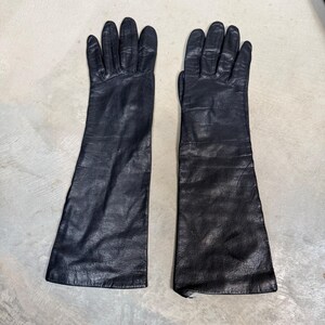 May include: A pair of long, black leather gloves featuring a smooth texture, five fingers, and a wide cuff that extends past the wrist. The gloves have visible stitching along the fingers and a natural grain pattern on the surface of the material.