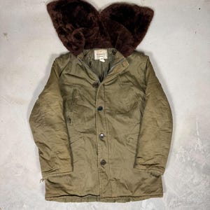 May include: An olive green parka featuring a large, dark brown faux fur hood, a front zip fastening, and four dark buttons. The jacket includes two chest pockets and two lower pockets, with a Windward brand label visible at the inner neckline.