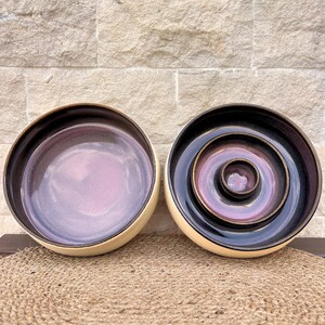 May include: Two ceramic bowls with a smooth, cream-colored exterior and a deep, glossy interior featuring a gradient of dark purple and soft lavender. The right bowl contains two smaller, nested circular dishes with the same color scheme.