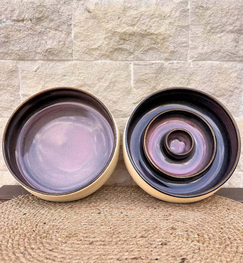 May include: Two ceramic bowls with a smooth, cream-colored exterior and a deep, glossy interior featuring a gradient of dark purple and soft lavender. The right bowl contains two smaller, nested circular dishes with the same color scheme.