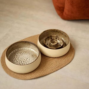 May include: Two round ceramic bowls with cream-colored outer walls and dark, speckled brown interiors. One bowl features a flat base, while the other contains a central raised circular pillar. The bowls are placed on an oval woven jute mat.