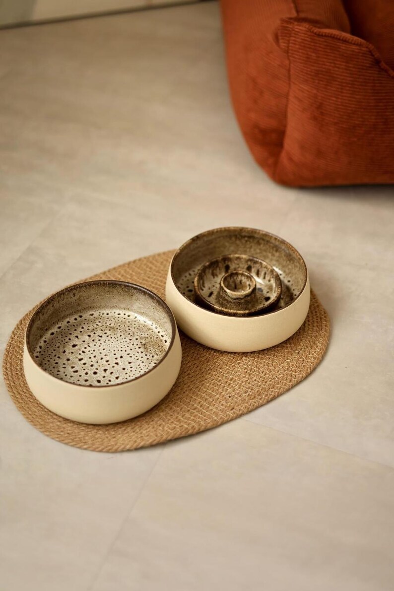 May include: Two round ceramic bowls with cream-colored outer walls and dark, speckled brown interiors. One bowl features a flat base, while the other contains a central raised circular pillar. The bowls are placed on an oval woven jute mat.