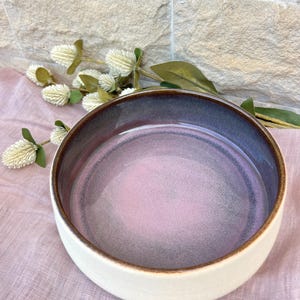 May include: A round ceramic bowl with a smooth, matte off-white exterior and a glossy interior featuring a gradient of soft pink transitioning into a deep purple rim. The bowl has a dark brown, exposed clay edge along the top circular rim.