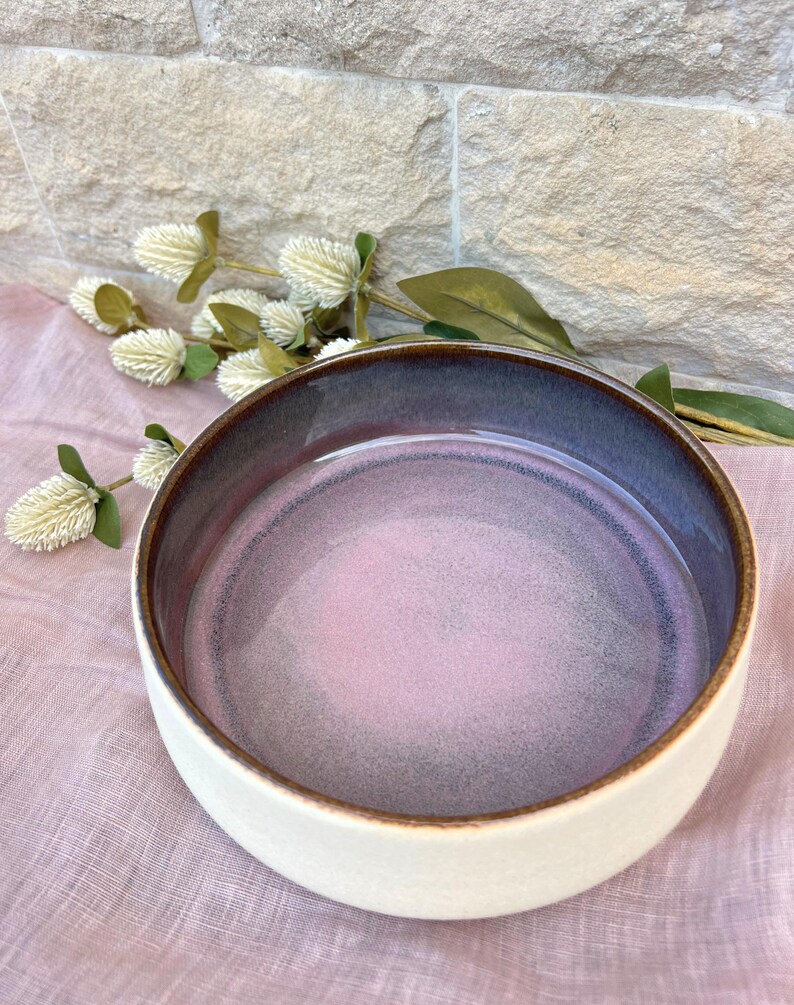 May include: A round ceramic bowl with a smooth, matte off-white exterior and a glossy interior featuring a gradient of soft pink transitioning into a deep purple rim. The bowl has a dark brown, exposed clay edge along the top circular rim.