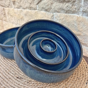 May include: A deep blue ceramic bowl featuring a raised concentric ring design in the center. The bowl has a smooth, glossy finish with dark brown edges along the rim and the interior rings, creating a distinct contrast against the blue glaze.