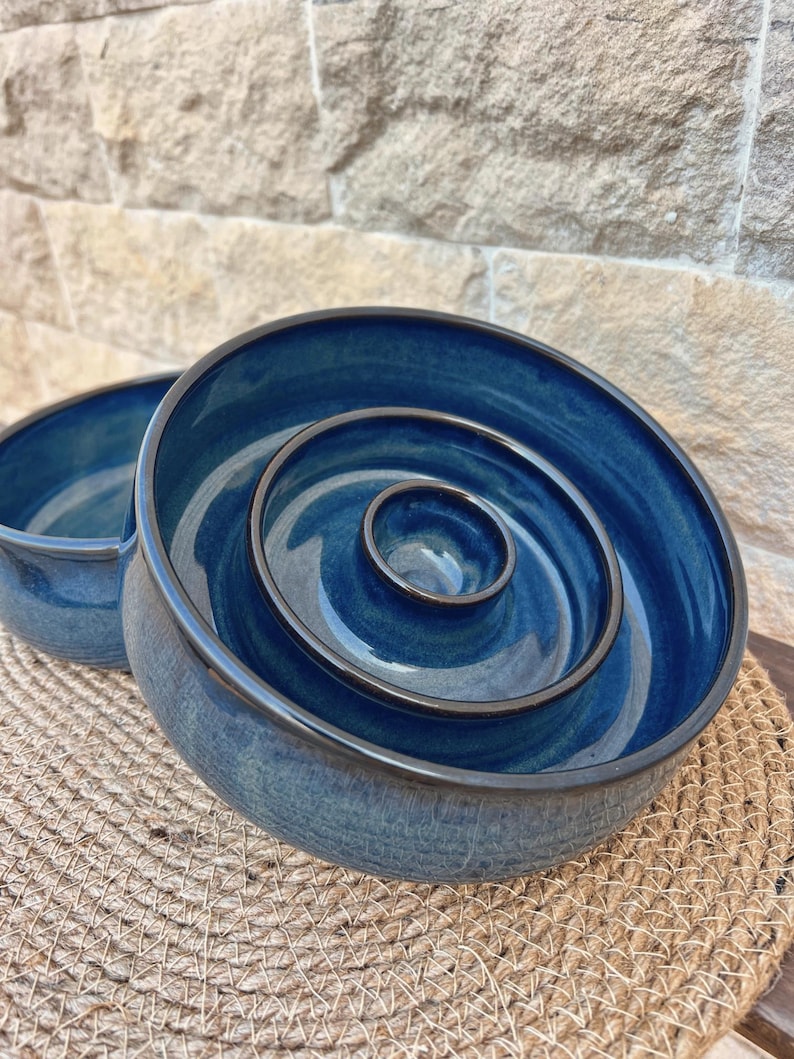 May include: A deep blue ceramic bowl featuring a raised concentric ring design in the center. The bowl has a smooth, glossy finish with dark brown edges along the rim and the interior rings, creating a distinct contrast against the blue glaze.