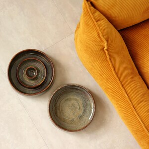 May include: Two round ceramic bowls with a dark, mottled glaze featuring shades of brown, gray, and green. One bowl is nested inside another, while the second bowl sits separately. A mustard yellow corduroy fabric is visible in the corner.