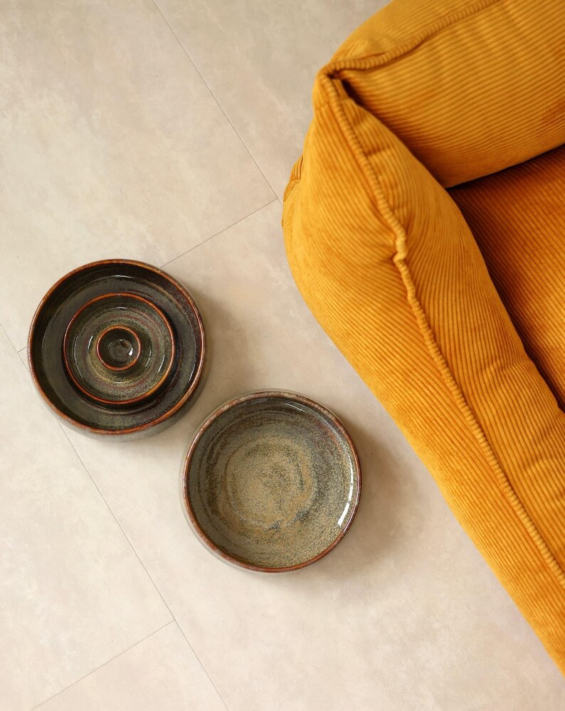 May include: Two round ceramic bowls with a dark, mottled glaze featuring shades of brown, gray, and green. One bowl is nested inside another, while the second bowl sits separately. A mustard yellow corduroy fabric is visible in the corner.