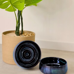May include: Two circular ceramic bowls with a deep, glossy, dark blue and purple iridescent glaze. One bowl is positioned upright to show the interior concentric circular ridges, while the other is placed beside it, showing the smooth exterior.