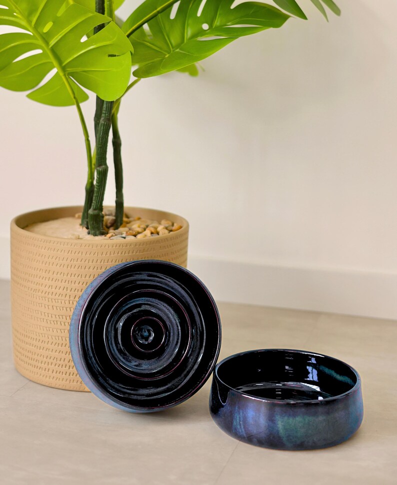 May include: Two circular ceramic bowls with a deep, glossy, dark blue and purple iridescent glaze. One bowl is positioned upright to show the interior concentric circular ridges, while the other is placed beside it, showing the smooth exterior.