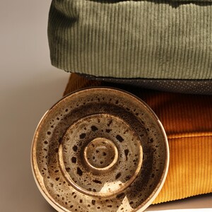May include: A round ceramic bowl with a speckled brown and cream glaze featuring concentric circular ridges on the interior surface, positioned in front of a stack of two corduroy cushions in sage green and mustard yellow colors.