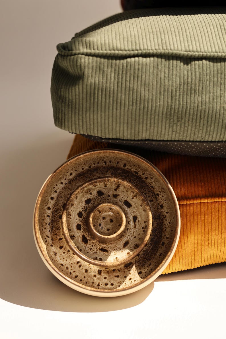 May include: A round ceramic bowl with a speckled brown and cream glaze featuring concentric circular ridges on the interior surface, positioned in front of a stack of two corduroy cushions in sage green and mustard yellow colors.