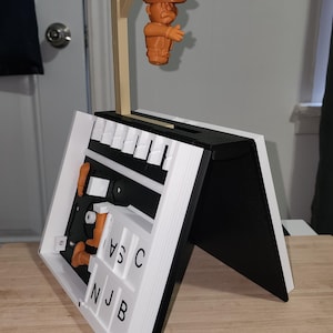 Puede incluir: Una figura de vaquero naranja impresa en 3D cuelga de una horca de madera sobre un tablero de juego blanco y negro. El tablero tiene una serie de teclas blancas y las letras "N", "J", "B", "V", "S" y "C".