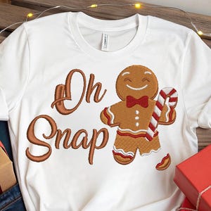 May include: White t-shirt with a gingerbread man design holding a candy cane. The words "Oh Snap" are embroidered in brown. The image includes wrapped red gifts and pine cones, suggesting a festive Christmas theme.
