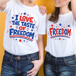Puede incluir: Dos camisetas blancas con texto gráfico en rojo y azul y detalles de estrellas. La camiseta de la izquierda muestra el texto I LOVE THE TASTE OF FREEDOM, mientras que la de la derecha muestra FREEDOM. Ambos diseños incluyen líneas curvas.