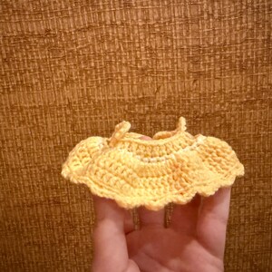 May include: A small, handmade, yellow crochet top with a scalloped edge. The top is held in a hand against a textured brown background. The crochet work is detailed and appears to be made of soft yarn.