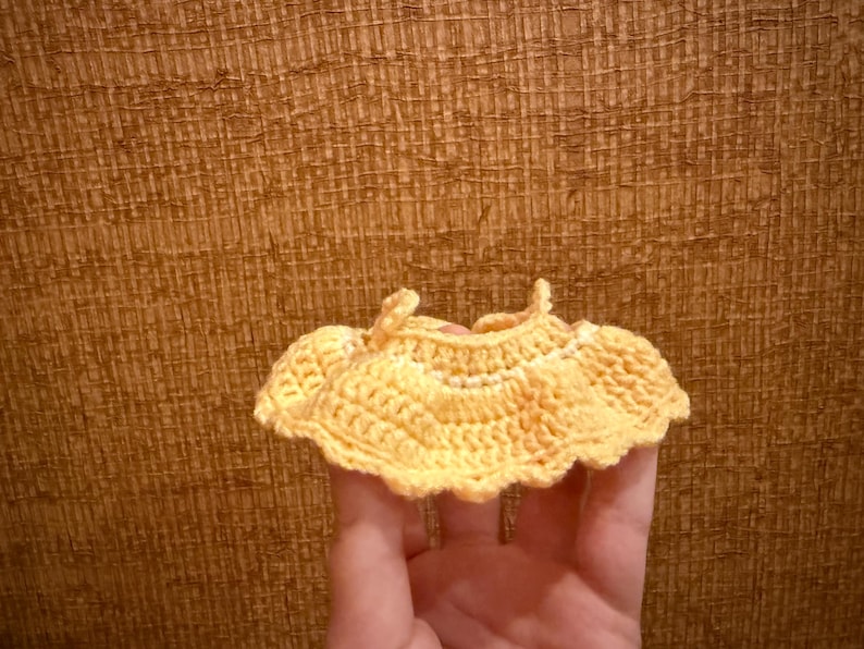 May include: A small, handmade, yellow crochet top with a scalloped edge. The top is held in a hand against a textured brown background. The crochet work is detailed and appears to be made of soft yarn.