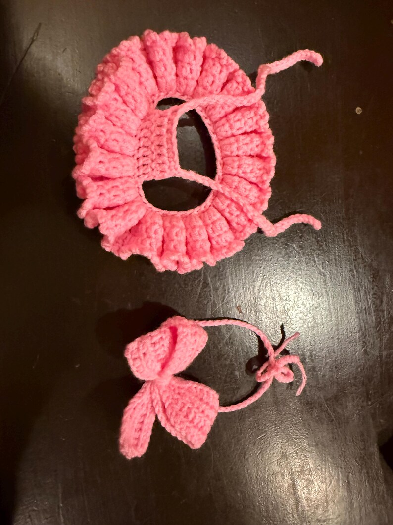 May include: A handmade, pink crocheted baby bonnet with a ruffled edge and attached ties. A matching pink crocheted bow is also included. The bonnet and bow are made of soft yarn, perfect for a newborn.
