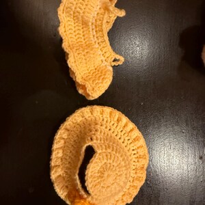 May include: Two handmade, crocheted items in a light orange color are displayed against a dark background. One item is a spiral shape, and the other is a curved shape. Both items have a textured, handmade appearance.