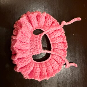 May include: A handmade, pink crocheted item with a ruffled edge and a central opening. The item has a circular shape with a textured, ribbed pattern. Two pink yarn ties extend from the side.
