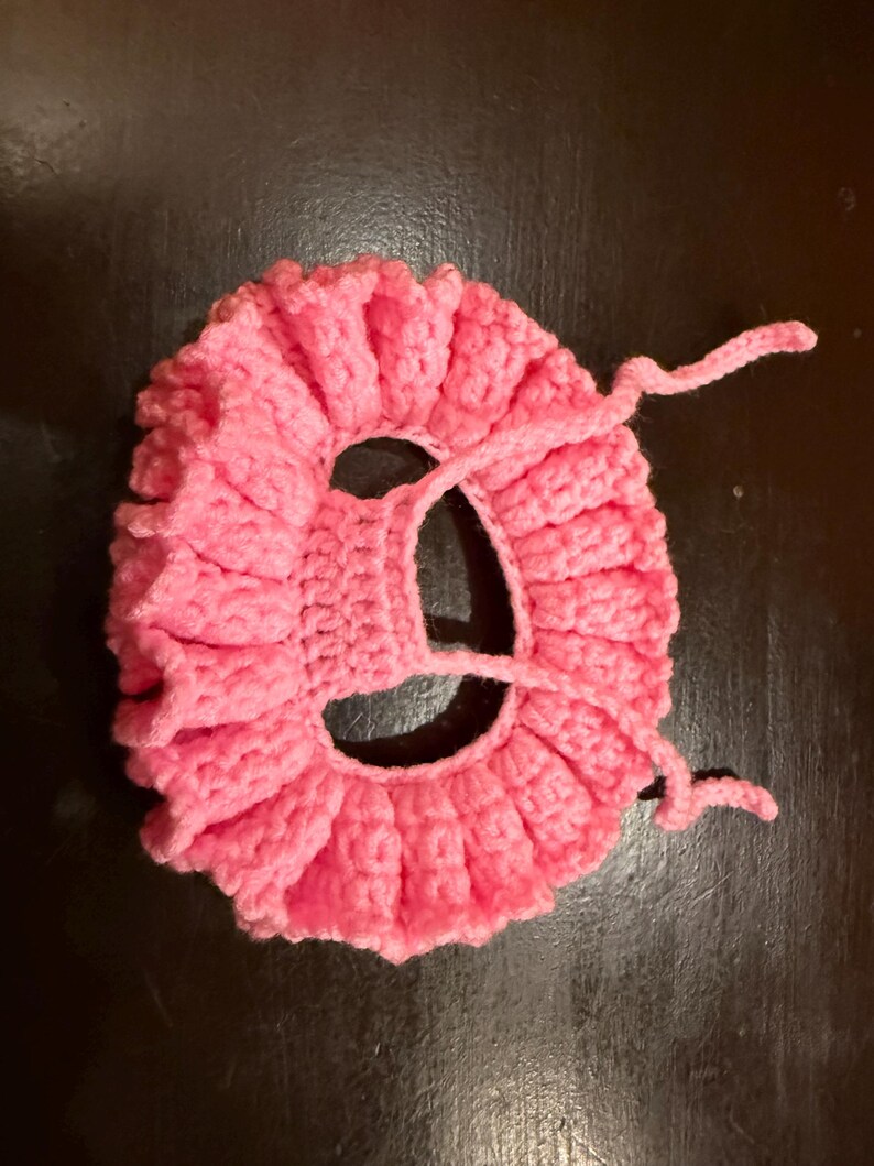 May include: A handmade, pink crocheted item with a ruffled edge and a central opening. The item has a circular shape with a textured, ribbed pattern. Two pink yarn ties extend from the side.