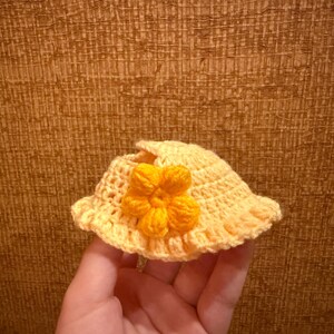 May include: A small, handmade, yellow crochet hat with a bright orange flower accent. The hat has a wide brim and is held in a hand, showcasing the intricate stitch work and sunny color palette.