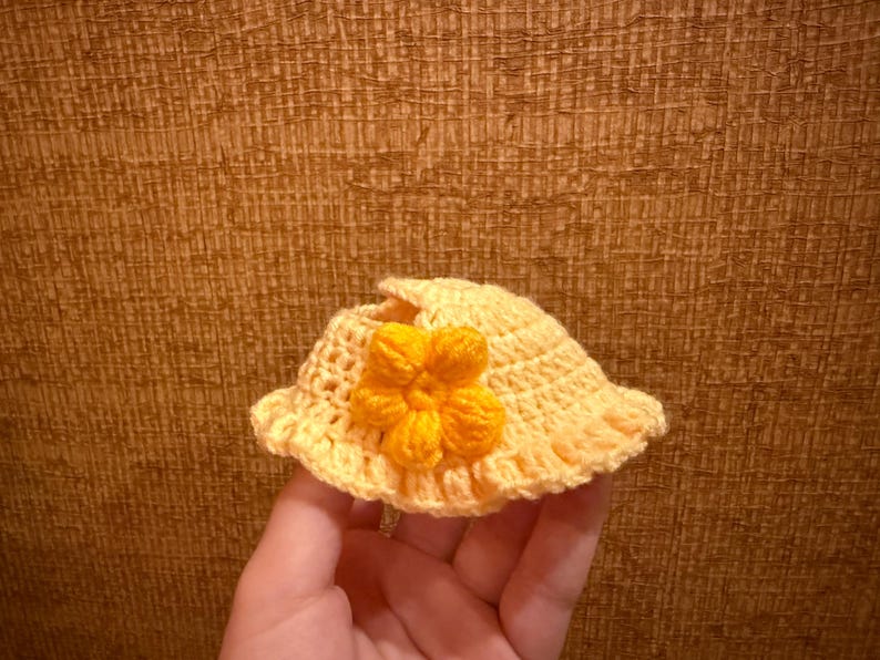 May include: A small, handmade, yellow crochet hat with a bright orange flower accent. The hat has a wide brim and is held in a hand, showcasing the intricate stitch work and sunny color palette.