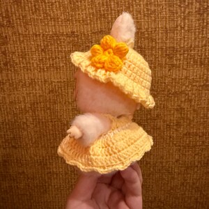May include: A handmade plush bunny wearing a yellow crocheted hat with a flower and a matching dress. The bunny is light pink with white fur accents. The hat and dress are a bright yellow color.