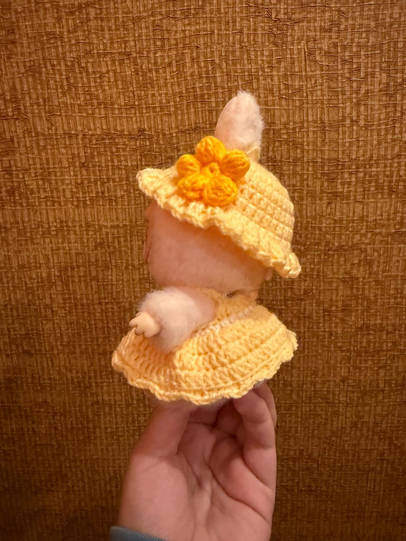 May include: A handmade plush bunny wearing a yellow crocheted hat with a flower and a matching dress. The bunny is light pink with white fur accents. The hat and dress are a bright yellow color.
