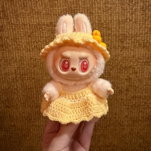 May include: A plush toy with a rabbit-like appearance, wearing a yellow crocheted dress and hat. The toy has a round face with red eyes and a neutral expression. The hat is adorned with a yellow flower.