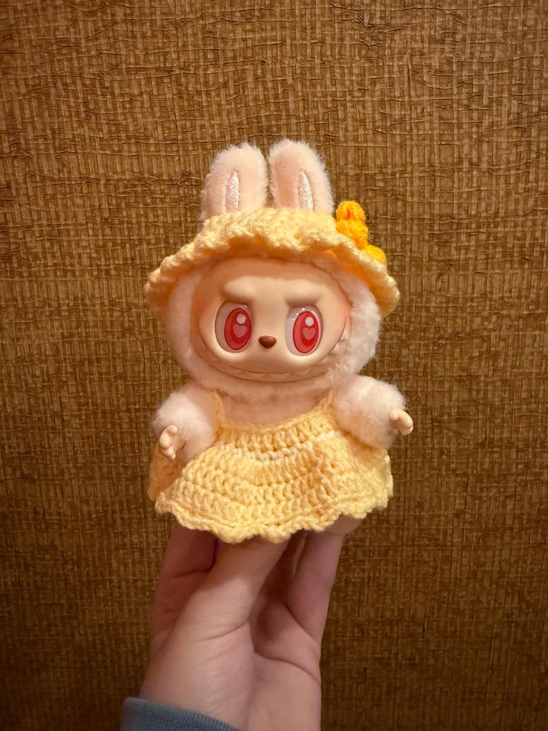 May include: A plush toy with a rabbit-like appearance, wearing a yellow crocheted dress and hat. The toy has a round face with red eyes and a neutral expression. The hat is adorned with a yellow flower.