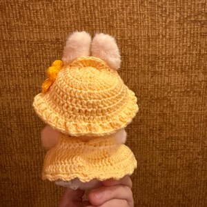May include: A handmade, yellow crochet doll wearing a matching hat with a yellow flower and a dress. The doll has white fluffy ears and a white body. The doll is held in a hand.