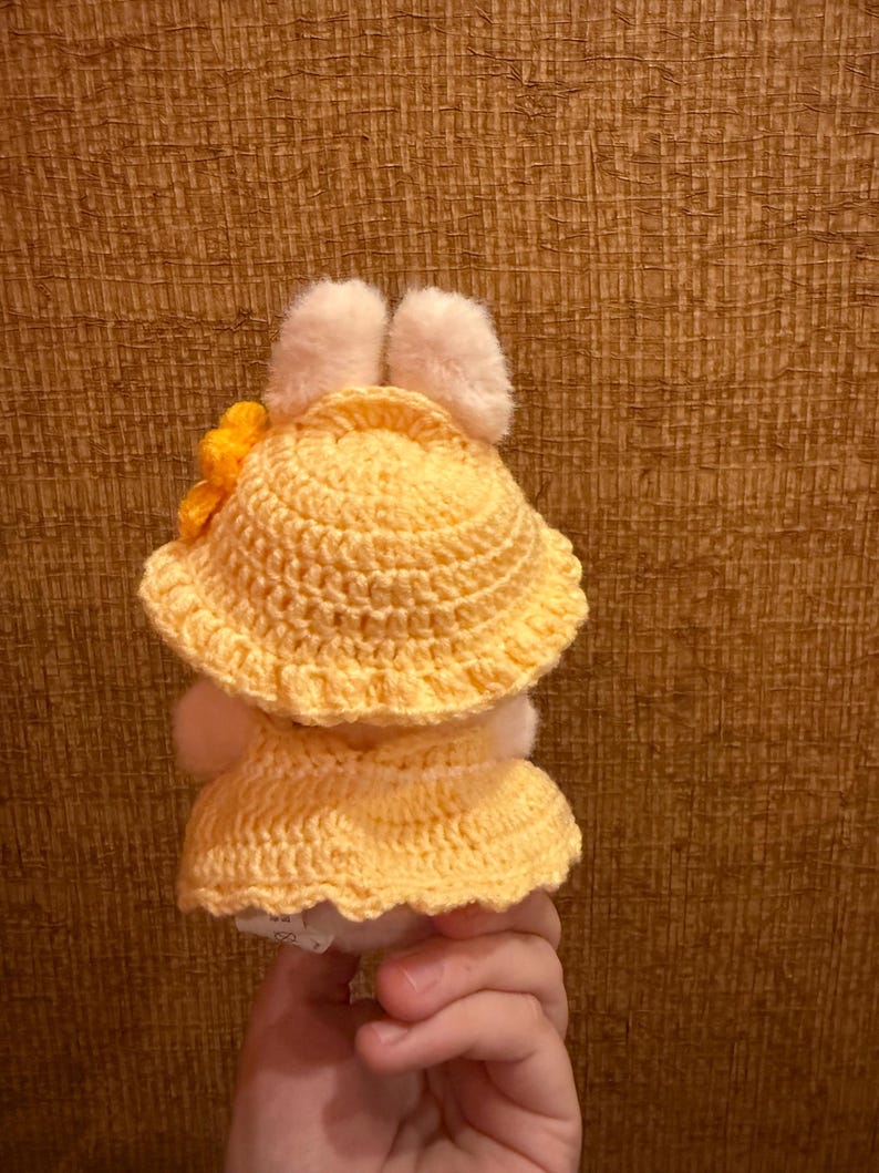 May include: A handmade, yellow crochet doll wearing a matching hat with a yellow flower and a dress. The doll has white fluffy ears and a white body. The doll is held in a hand.