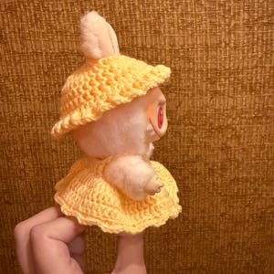 May include: A plush bunny doll wearing a yellow crocheted hat and dress. The doll has pink ears and red eyes. The hat has a wide brim, and the dress is a simple, flared style. The doll is held up by a hand.
