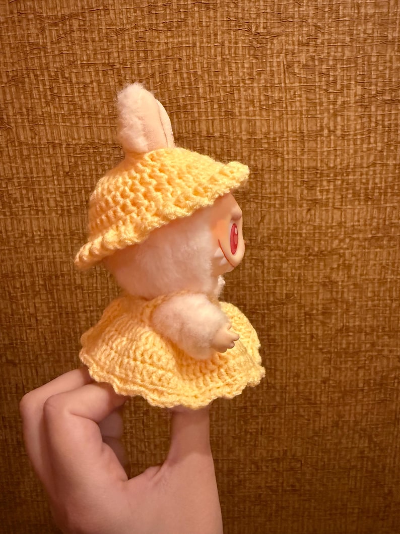 May include: A plush bunny doll wearing a yellow crocheted hat and dress. The doll has pink ears and red eyes. The hat has a wide brim, and the dress is a simple, flared style. The doll is held up by a hand.